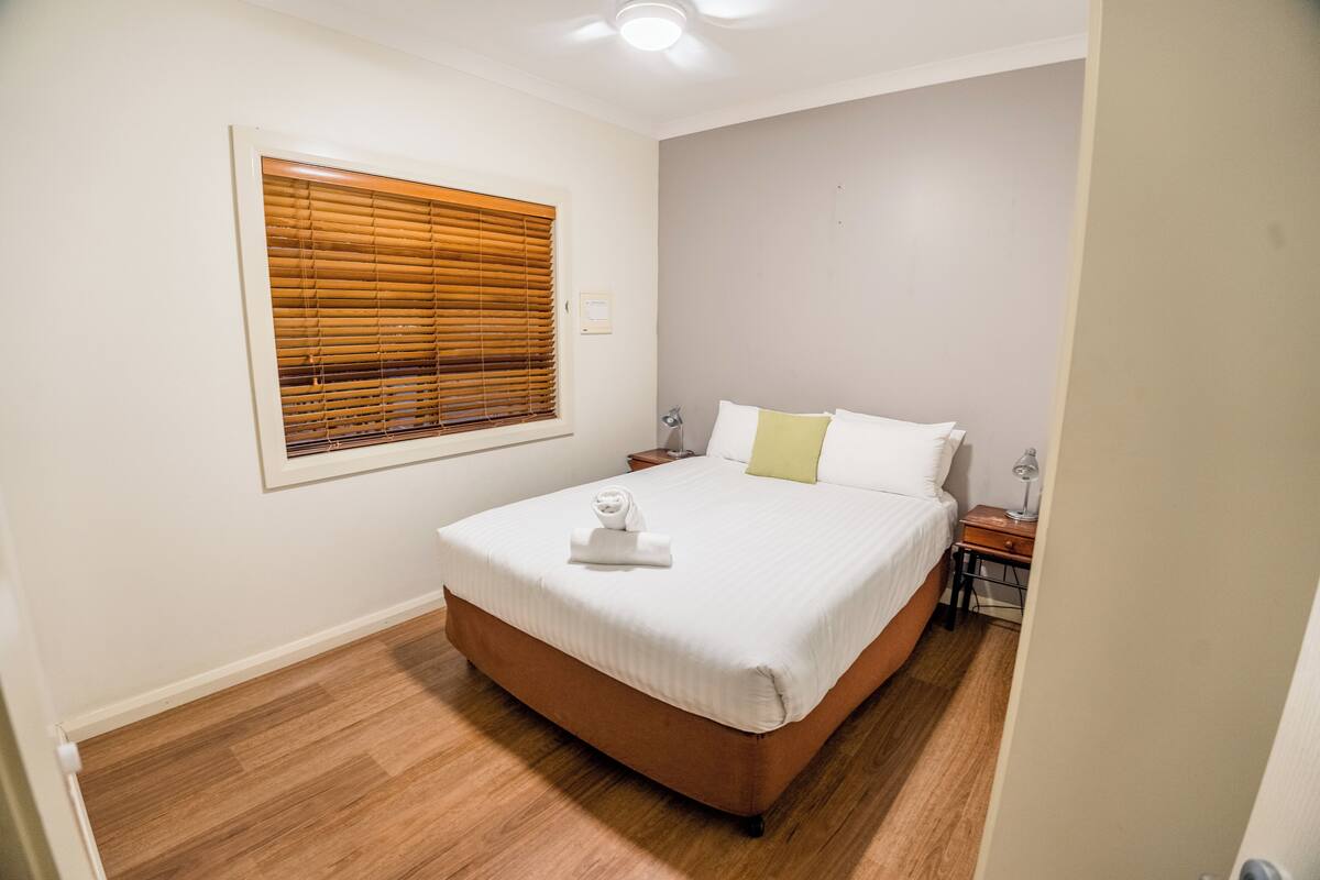 A bedroom with a bed, a window with blinds, a bedside table with a lamp, and a nightstand.
