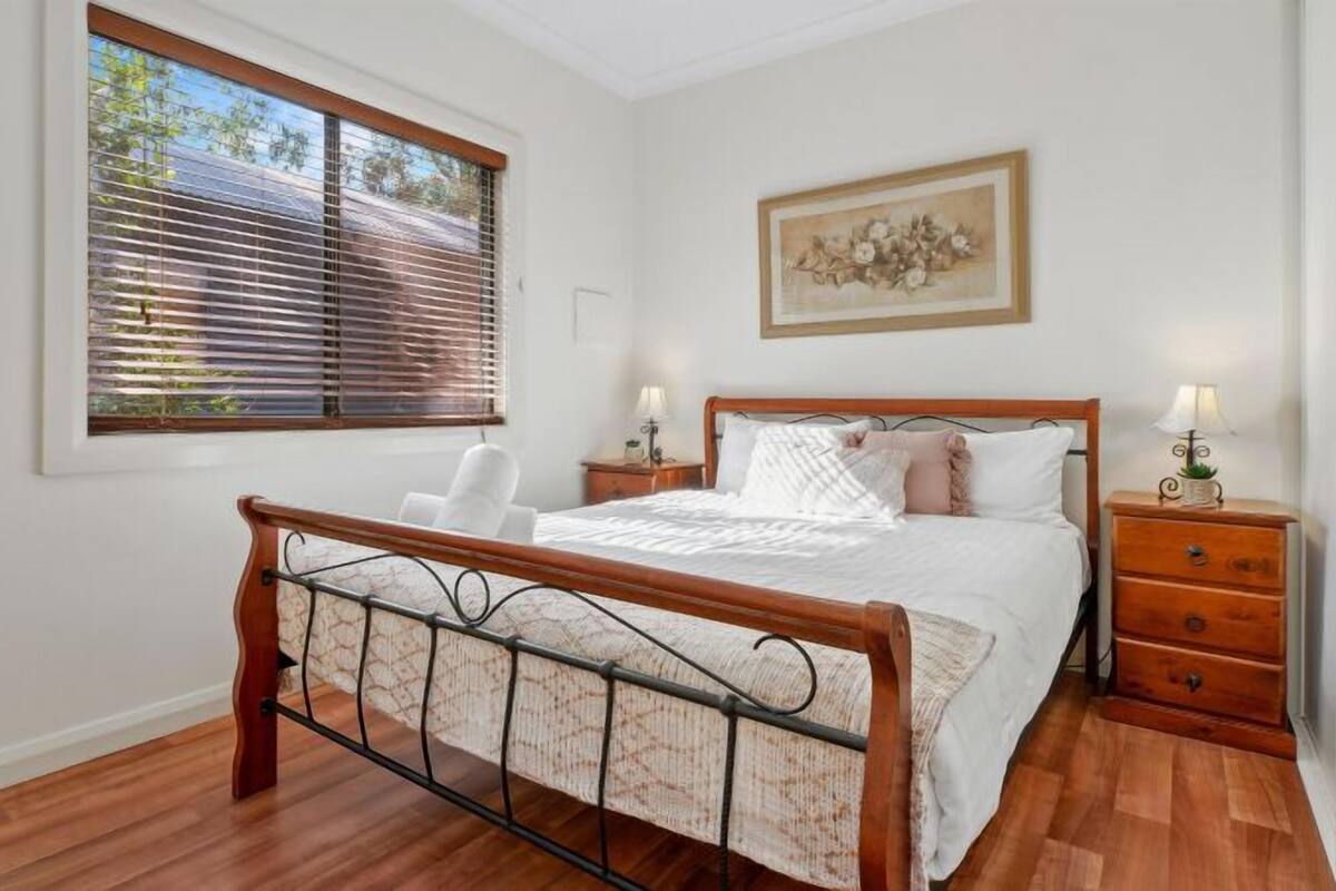 A bedroom with a wooden bed, a nightstand, a chair, a window with blinds, and a framed picture on the wall.