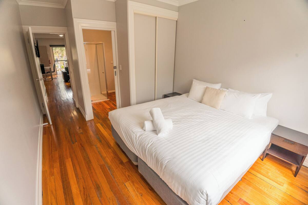 A bedroom with a large bed, wooden flooring, a wardrobe, and a nightstand.