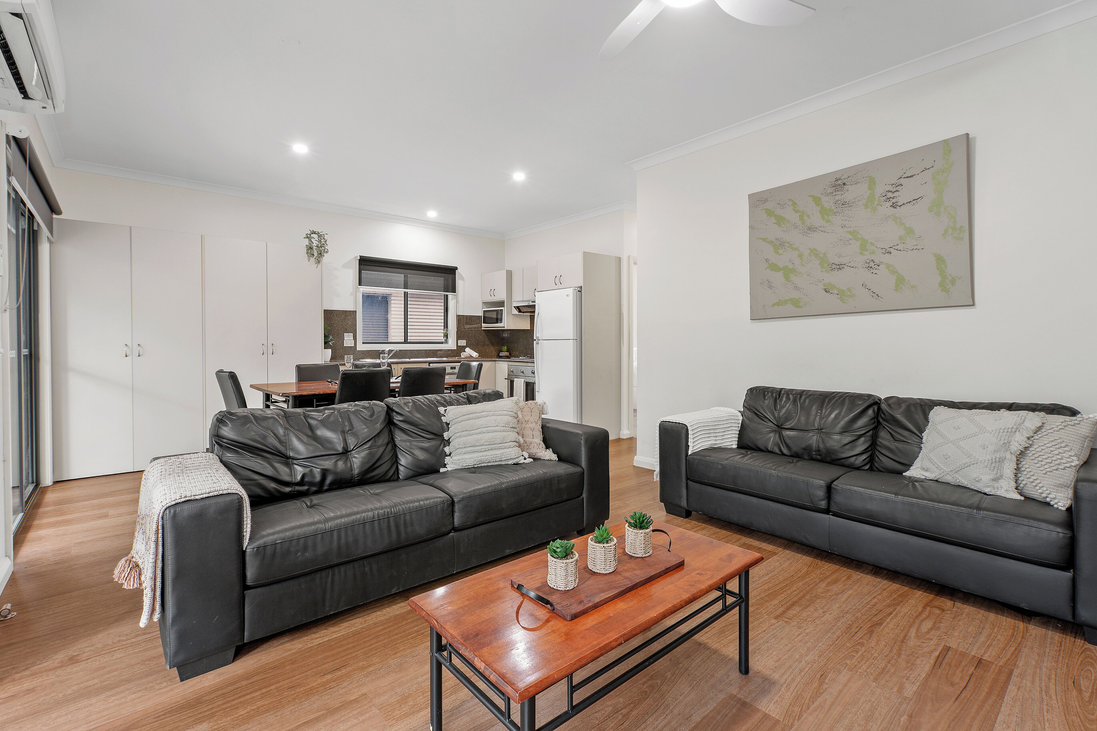 A modern living room with two black leather sofas, a wooden coffee table, and a kitchen in the background.