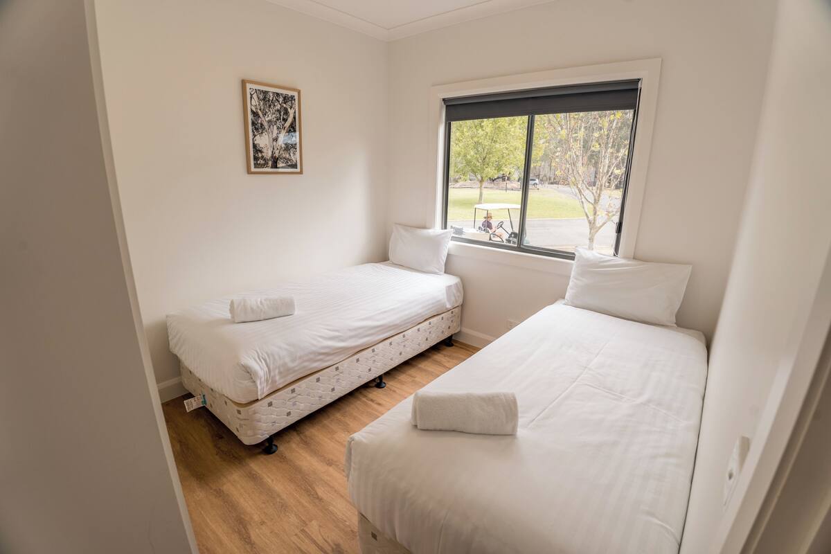 A room with two single beds, a window with a view of the outdoors, a framed picture on the wall, and a wooden floor.