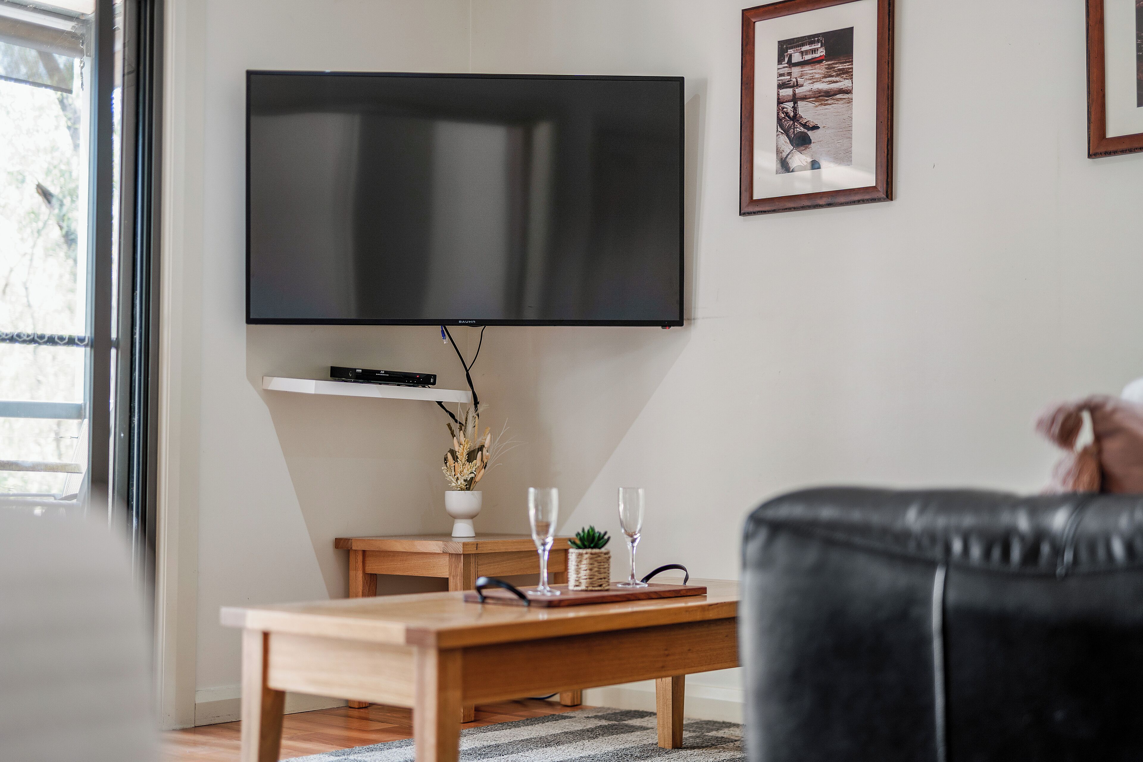 Una sala de estar con un televisor de pantalla plana montado en la pared, una mesa de centro de madera, un sofá de cuero negro y un cuadro enmarcado en la pared.