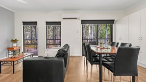 A modern living room with a black sofa, dining table, and chairs, large windows with a view of trees, and an air conditioning unit.