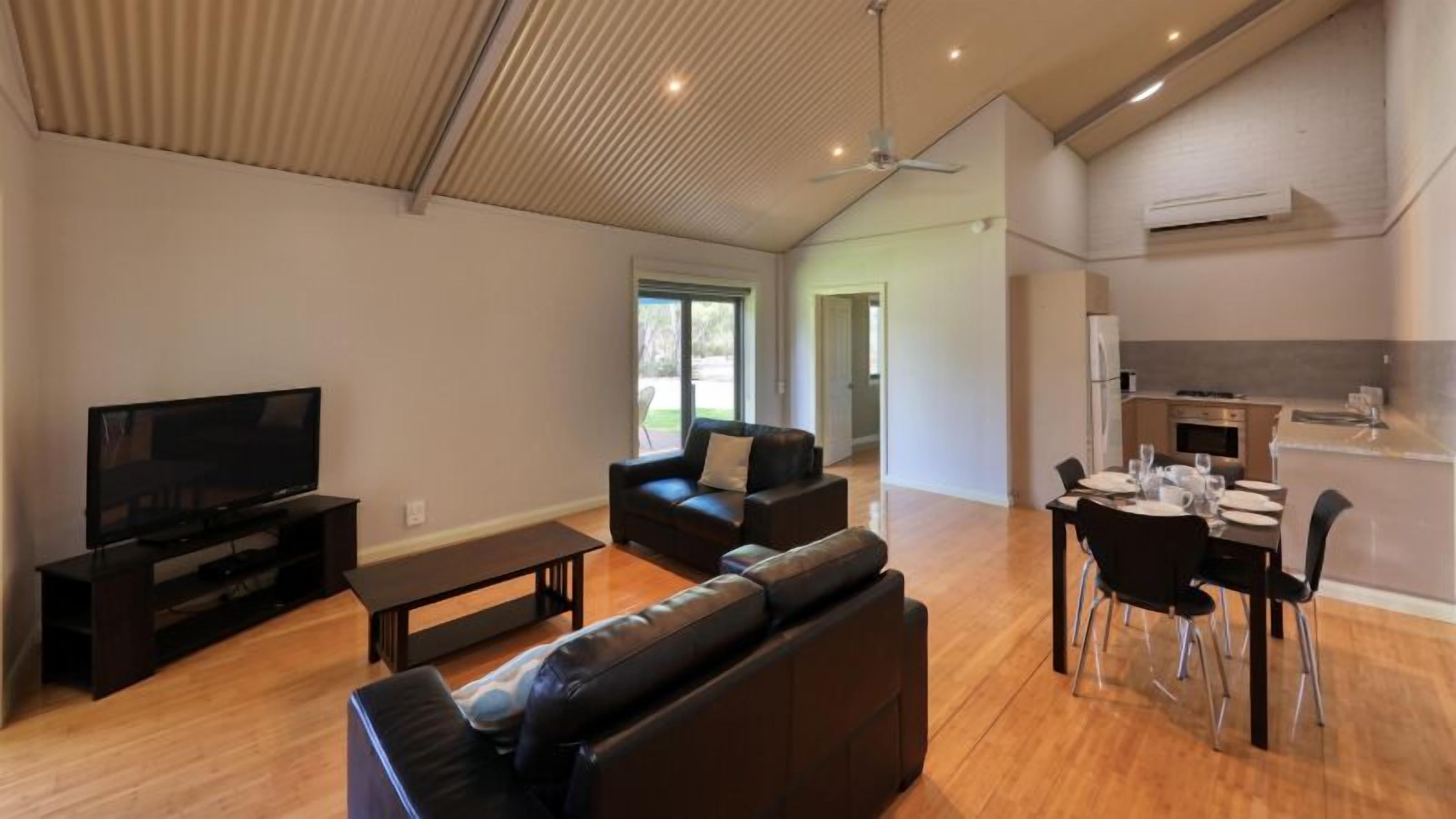 A spacious living area with a flat-screen TV, leather sofas, a dining table with chairs, and a kitchen area with a refrigerator and oven.