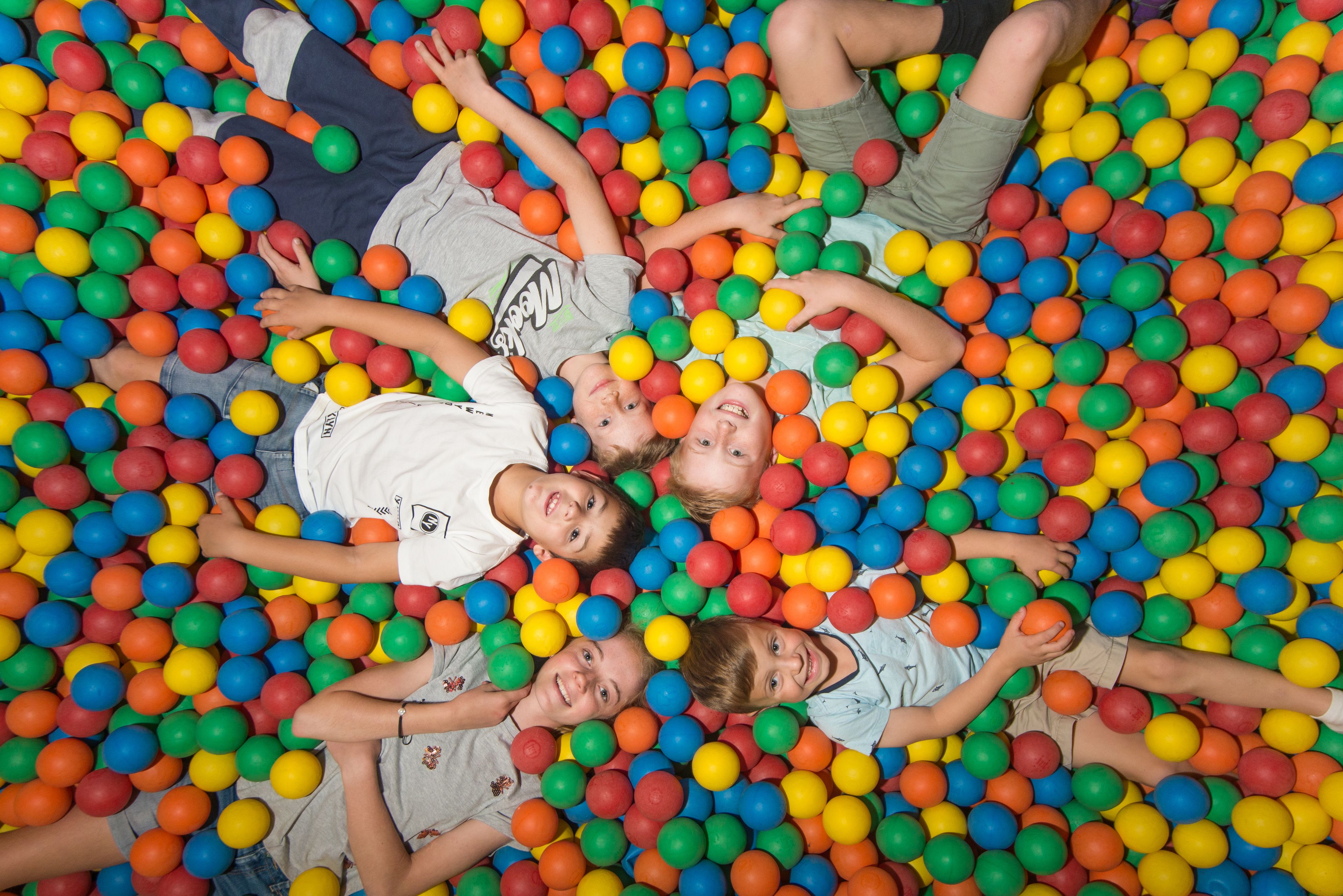 A group of children lying in a ball pit filled with multicolored plastic balls.