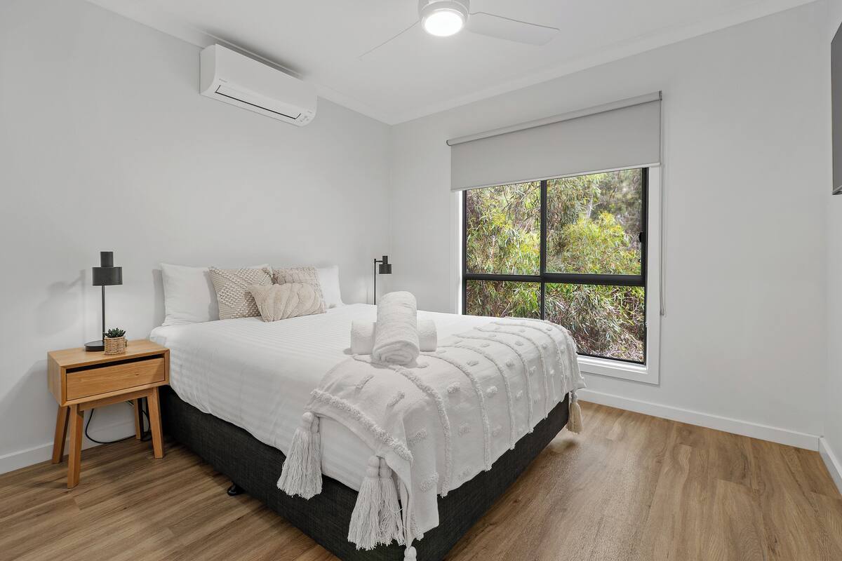 A bedroom with a bed, a nightstand, a window with a view of greenery, and an air conditioning unit.