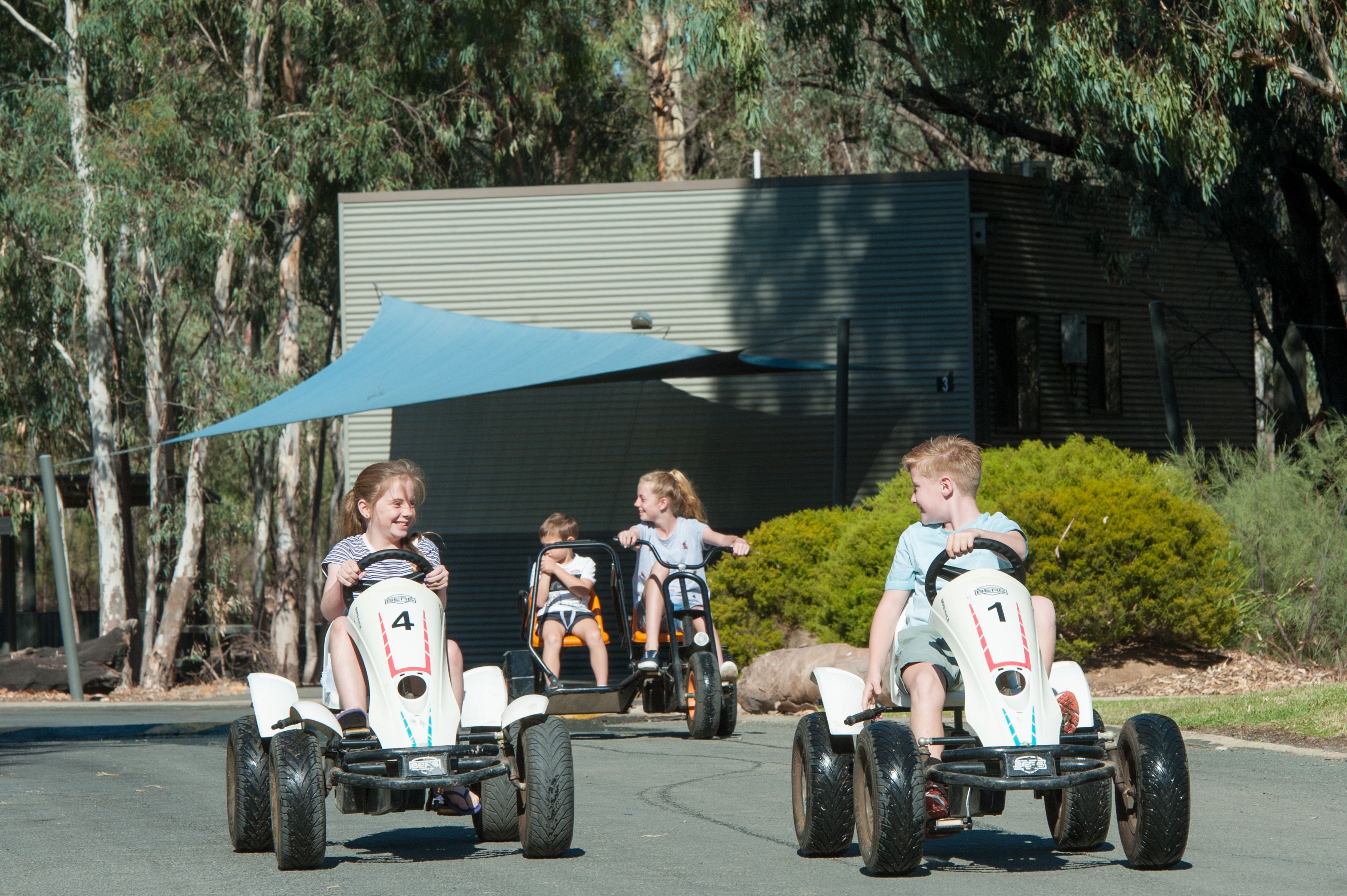 Four children riding go-karts on a paved road with a building and trees in the background.