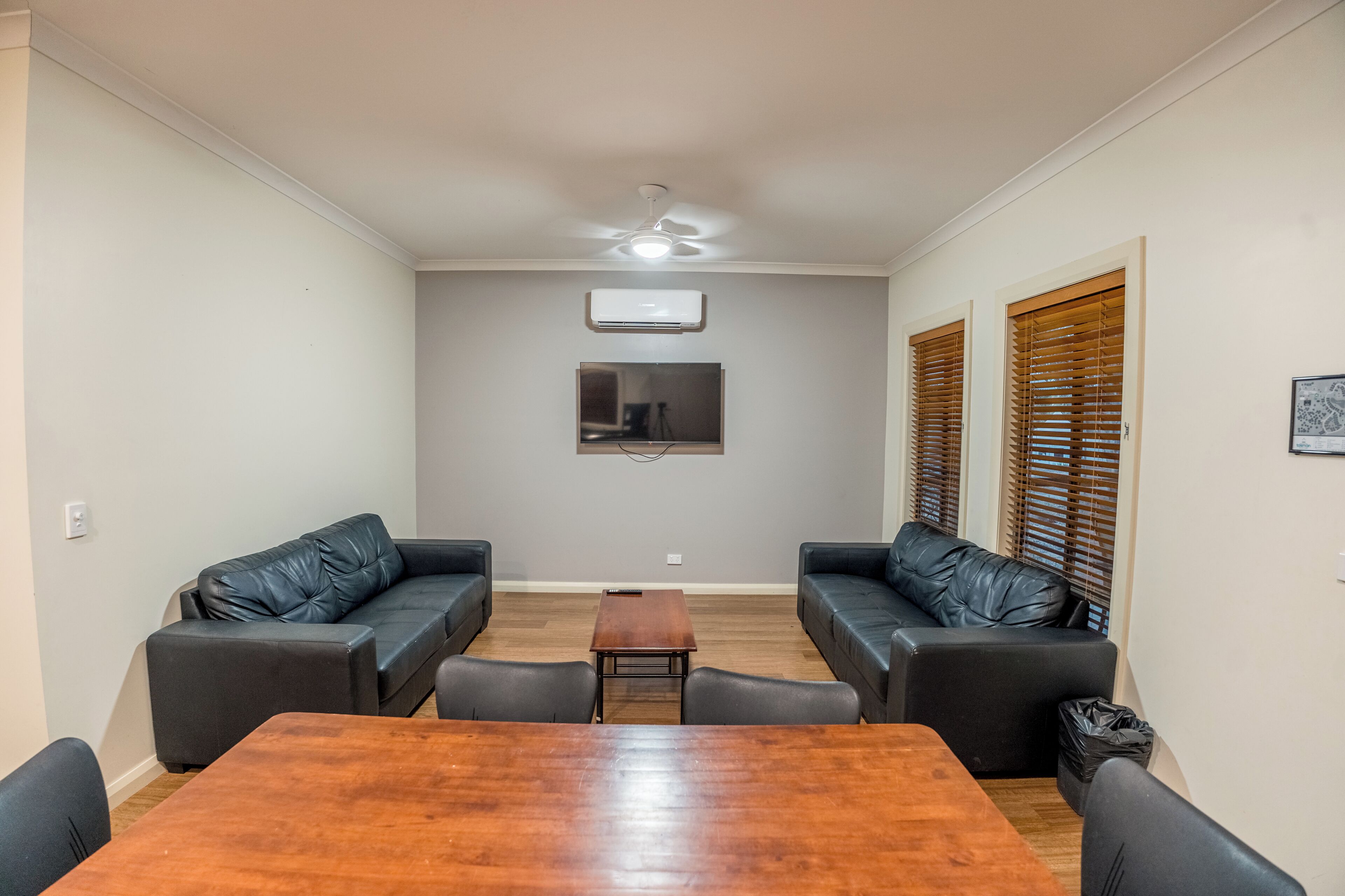 A living room with a wooden table, two black leather sofas, a ceiling fan, and a mounted air conditioner.