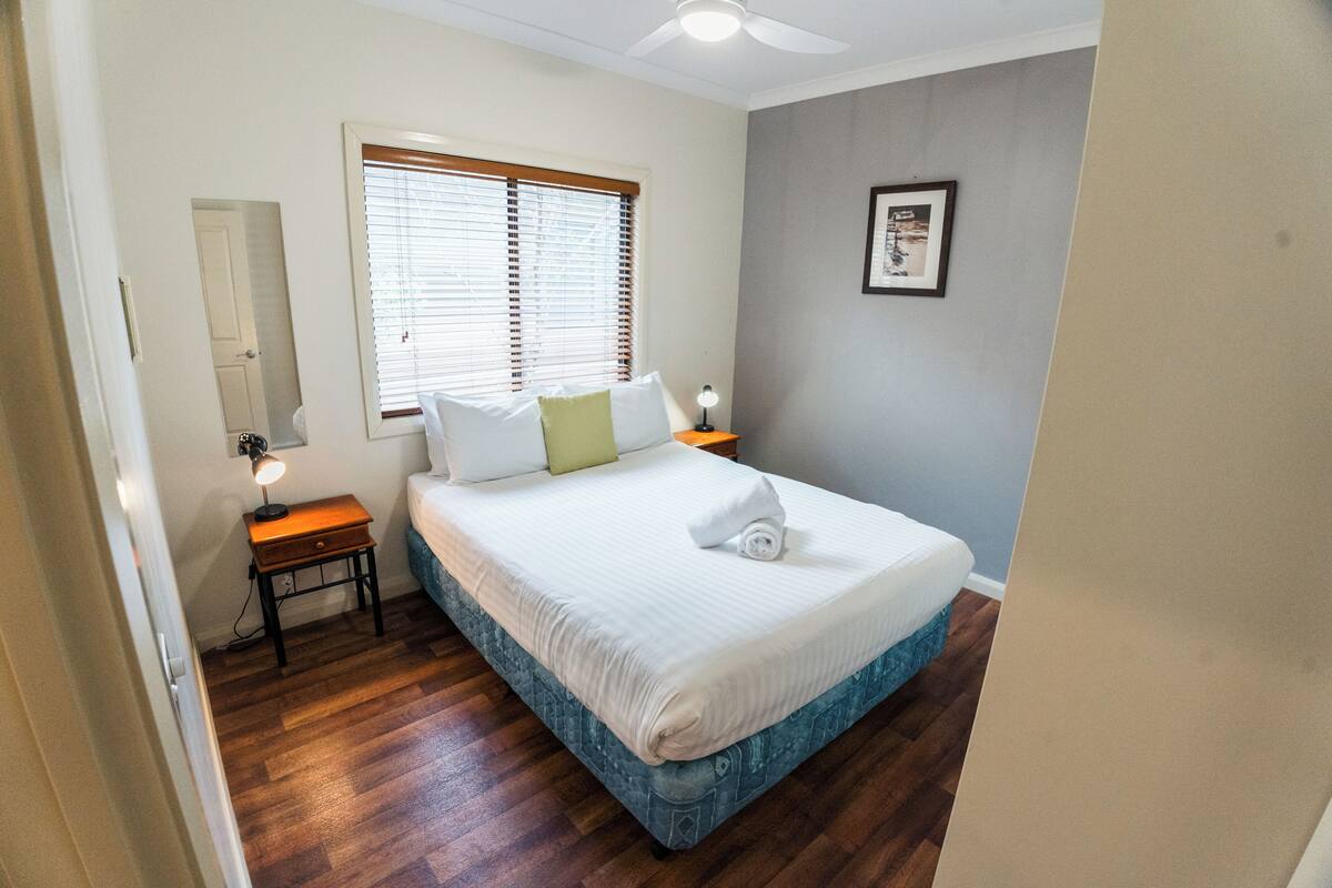 A bedroom with a bed, a nightstand with a lamp, a window with blinds, a door, and a framed picture on the wall.