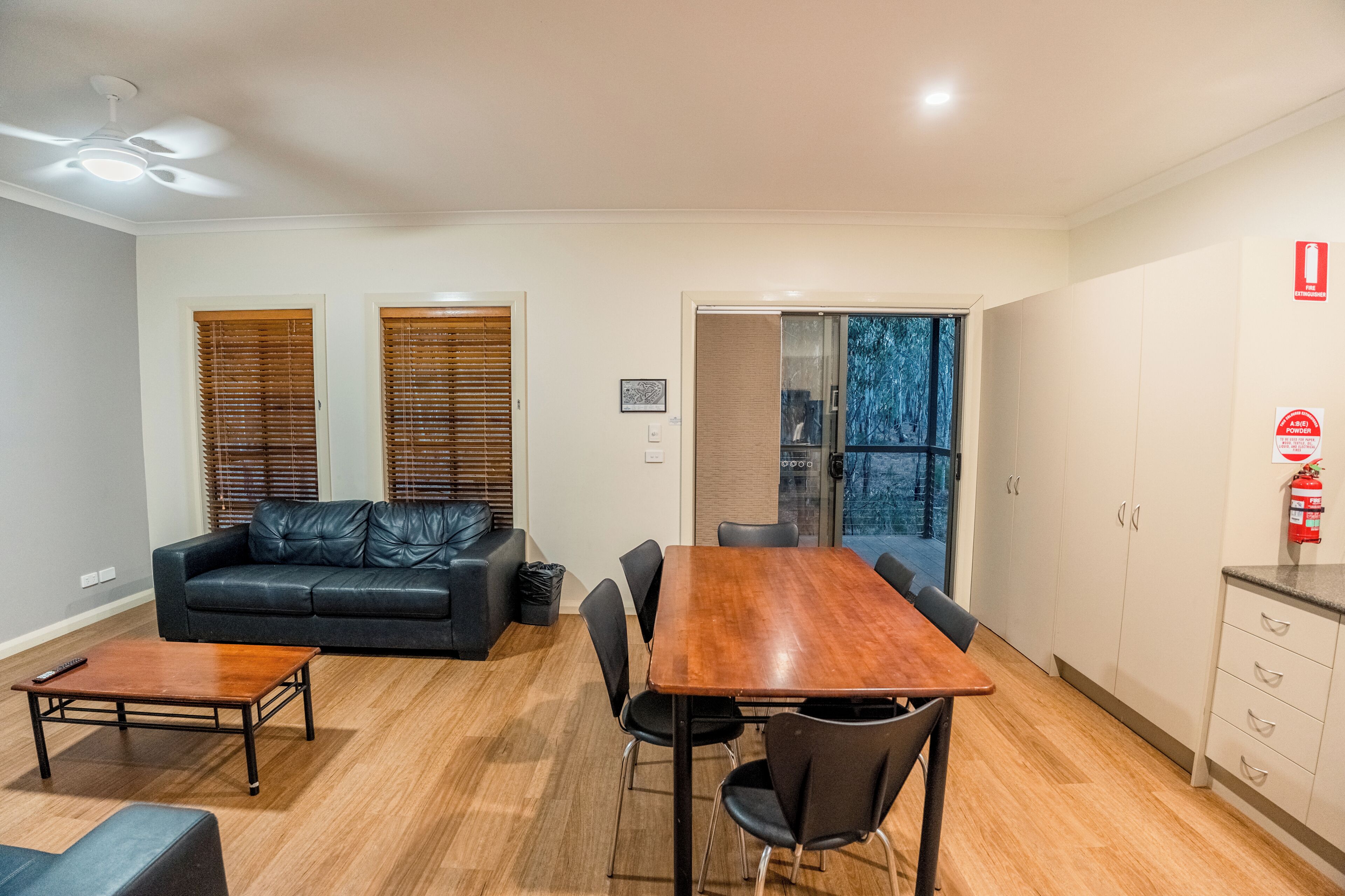 Una sala de estar moderna con un sofá de cuero negro, una mesa de centro de madera y una mesa de comedor con sillas. Hay una puerta de cristal que da a un balcón con vistas a árboles.