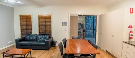 Una sala de estar moderna con un sofá de cuero negro, una mesa de centro de madera y una mesa de comedor con sillas. Hay una puerta de cristal que da a un balcón con vistas a árboles.