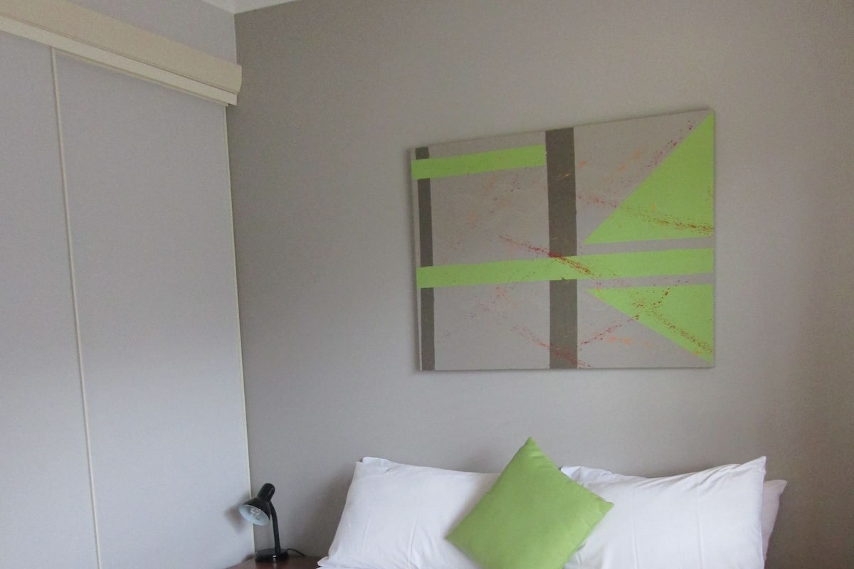 A neatly made bed with white linens and a green pillow, a white towel on the bed, and a painting on the wall.