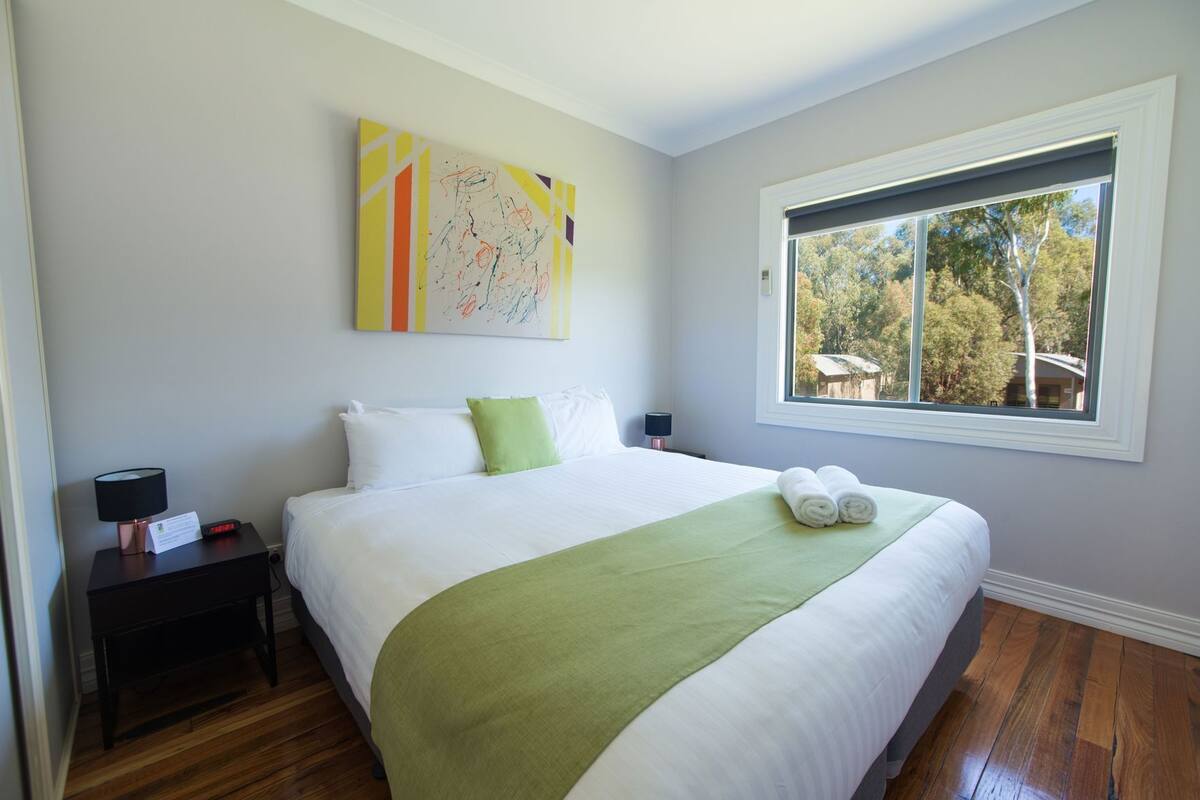 A hotel room with a large bed, a nightstand with a lamp, a painting on the wall, and a window with a view of trees.
