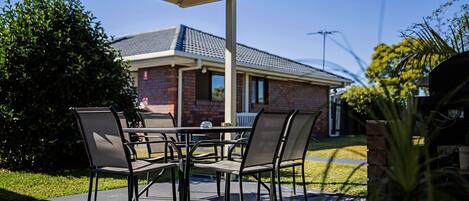 An outdoor patio with a table and chairs, a covered area, and a brick building in the background.