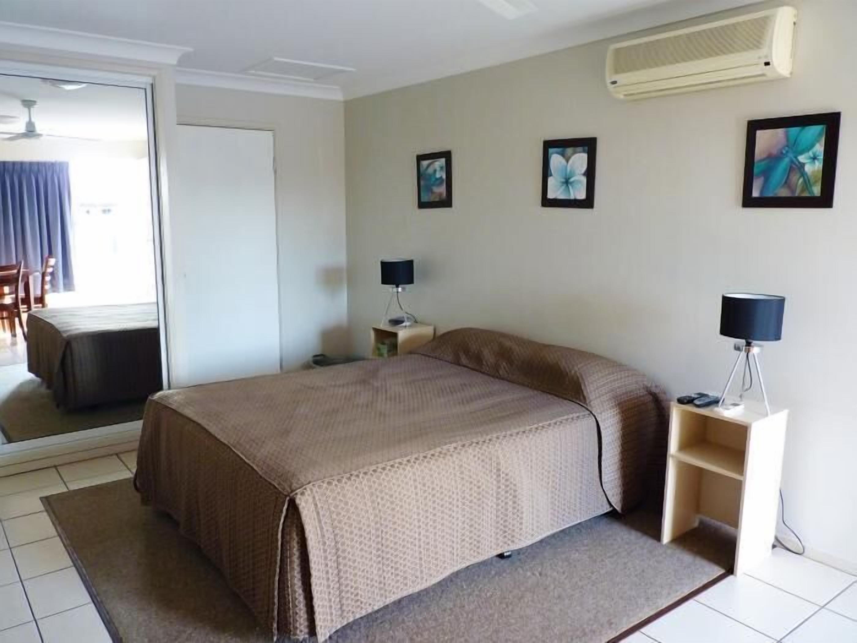 A hotel room with a large bed, a desk with a lamp, a chair, and a small table. There are three framed pictures on the wall and an air conditioning unit above the bed.