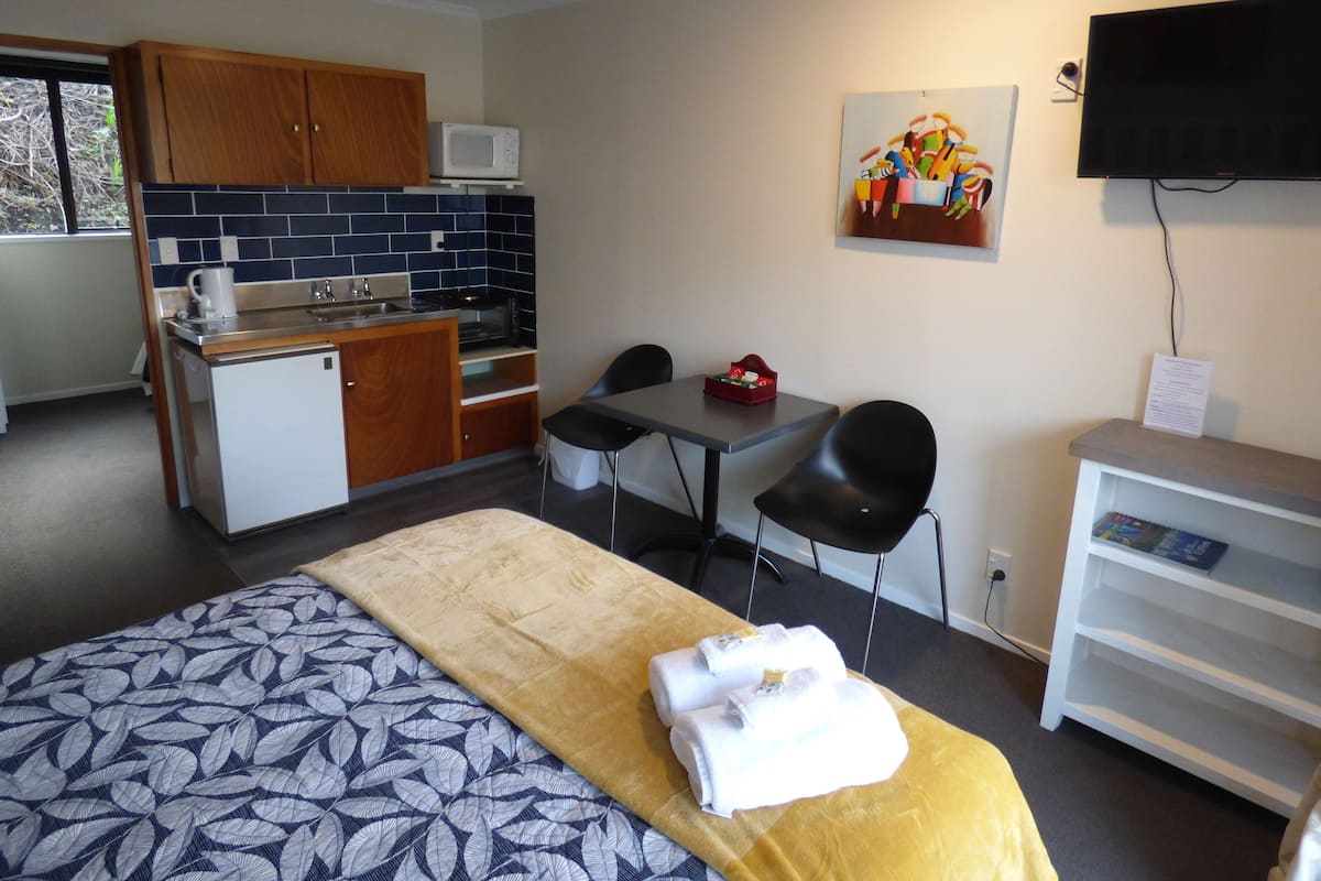 A compact hotel room with a bed, a small kitchenette, a dining area, and a TV.