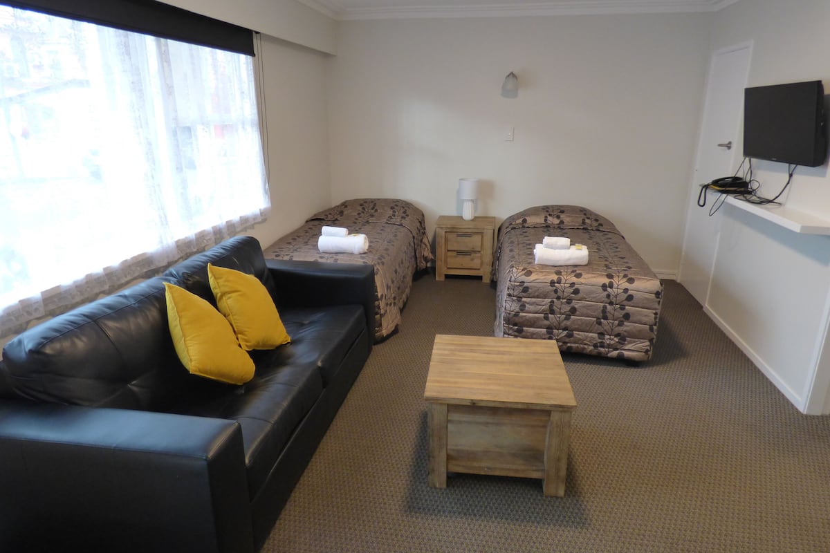 A room with a black leather sofa, two single beds with patterned bedding, a wooden nightstand, a small wooden table, and a television mounted on the wall.