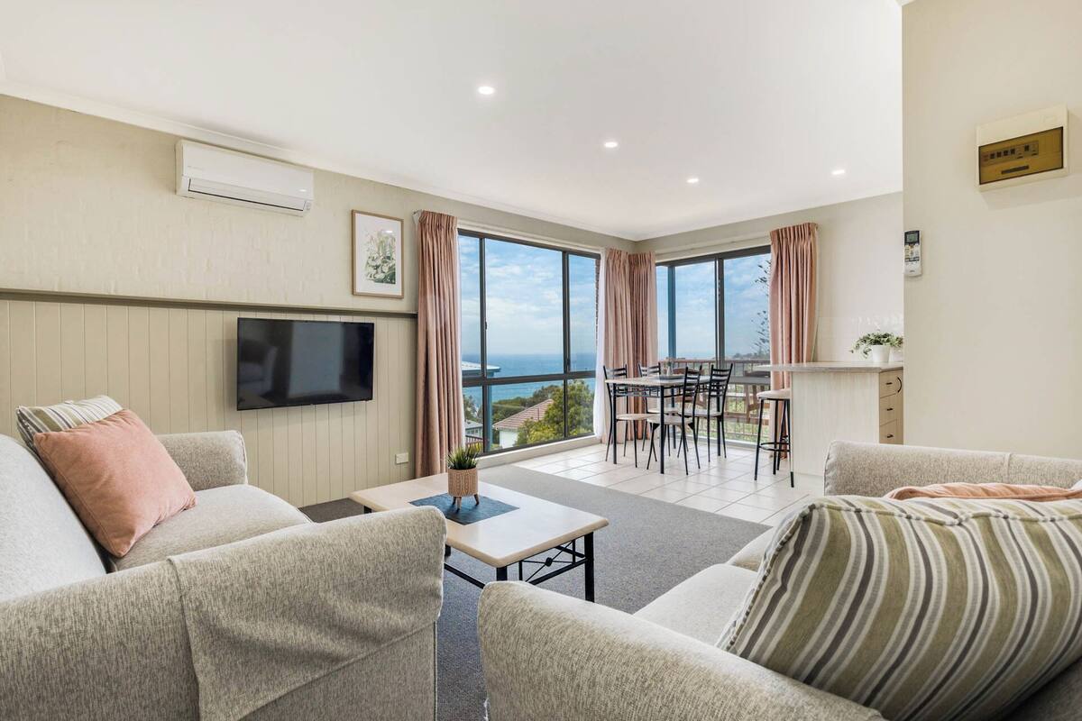 A modern living room with a flat-screen TV, a dining area with a table and chairs, and a balcony with a view of the sea.