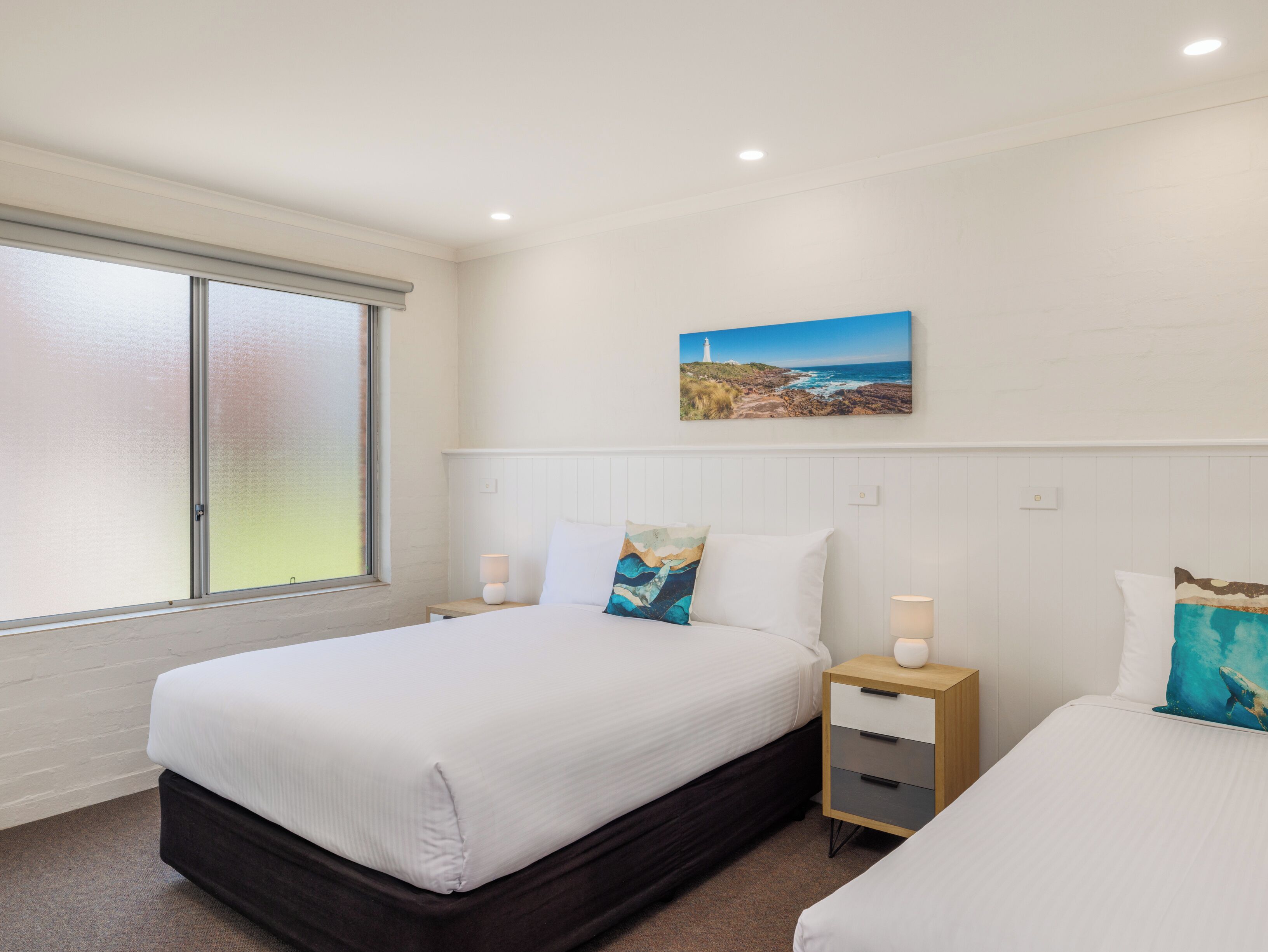 Une chambre à coucher avec un grand lit, une table de chevet, une lampe fixée au mur et un tableau représentant un paysage côtier.
