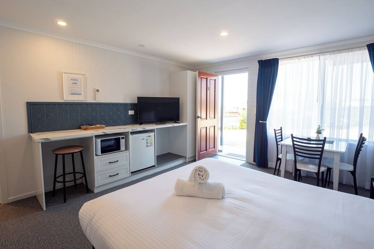 A hotel room with a bed, a desk with a TV, a microwave, and a small refrigerator. There is a window with curtains and a view of the outside.