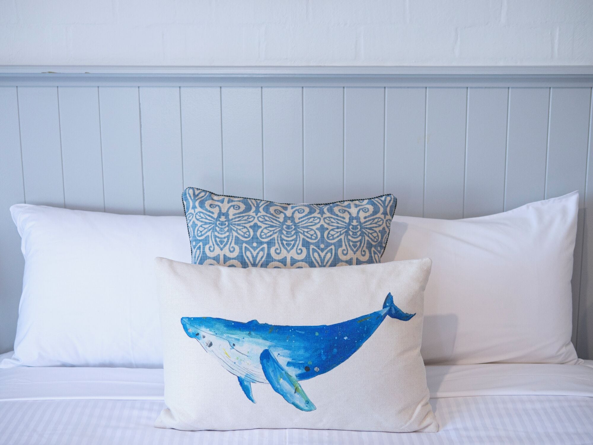 Ein Bett mit weißer Bettwäsche und zwei dekorativen Kissen, eines mit einem blauen Walmotiv und das andere mit blau-weißem Muster.
