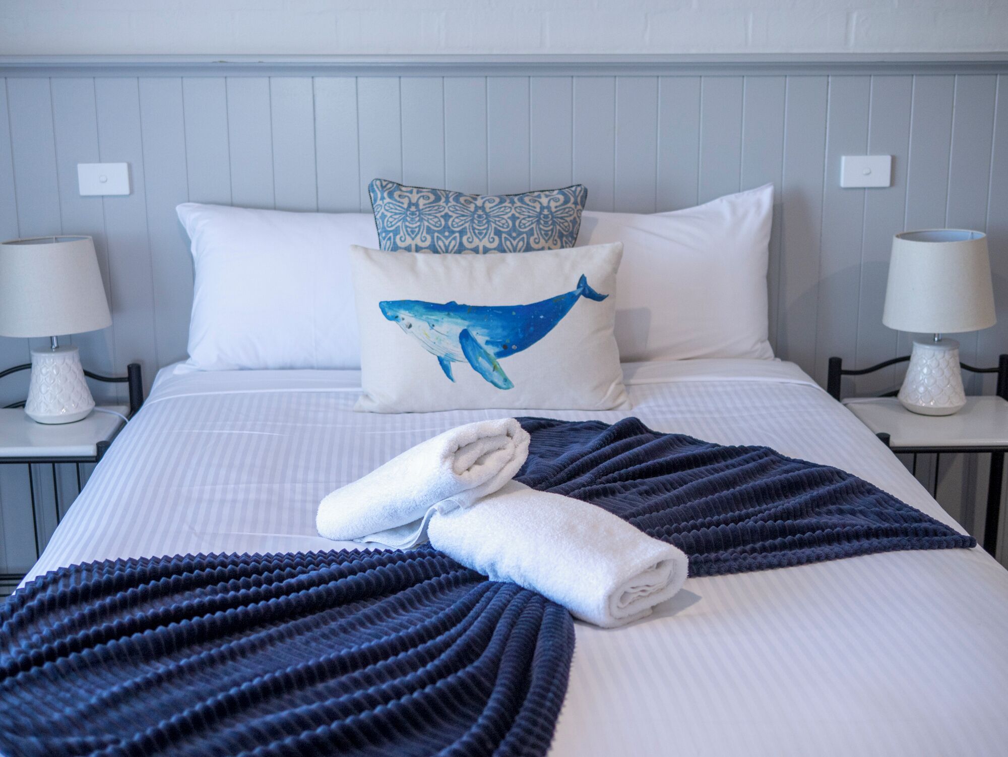 Un letto rifatto con un cuscino azzurro e bianco, un asciugamano piegato e una coperta blu scuro.