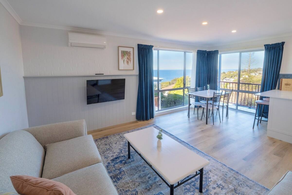 A modern living room with a flat-screen TV, a sofa, a dining area with a table and chairs, and a balcony with a view of the sea.