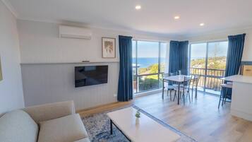 Una sala de estar moderna con una televisión de pantalla plana, un sofá, una zona de comedor con mesa y sillas, y un balcón con vista al mar.