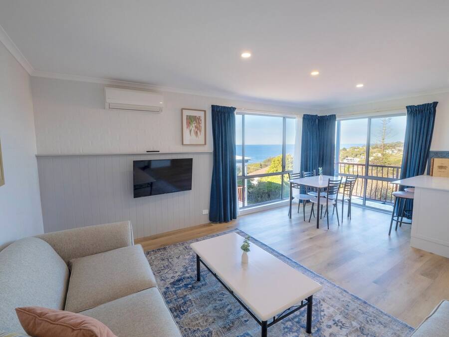 Una sala de estar moderna con una televisión de pantalla plana, un sofá, una zona de comedor con mesa y sillas, y un balcón con vista al mar.