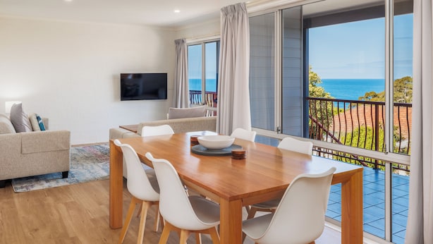 A spacious living room with a wooden dining table, white chairs, a flat-screen TV, and a balcony with a view of the sea. - Heritage House Motel & Units (Eden)