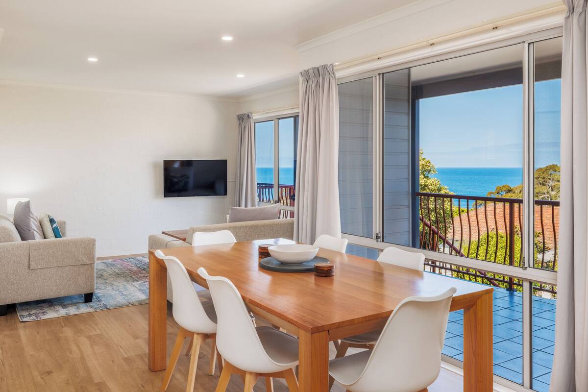 A spacious living room with a wooden dining table, white chairs, a flat-screen TV, and a balcony with a view of the sea.