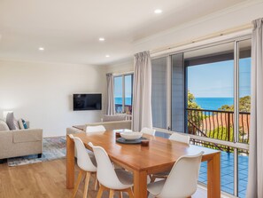 A spacious living room with a wooden dining table, white chairs, a flat-screen TV, and a balcony with a view of the sea. - Heritage House Motel & Units (Eden)