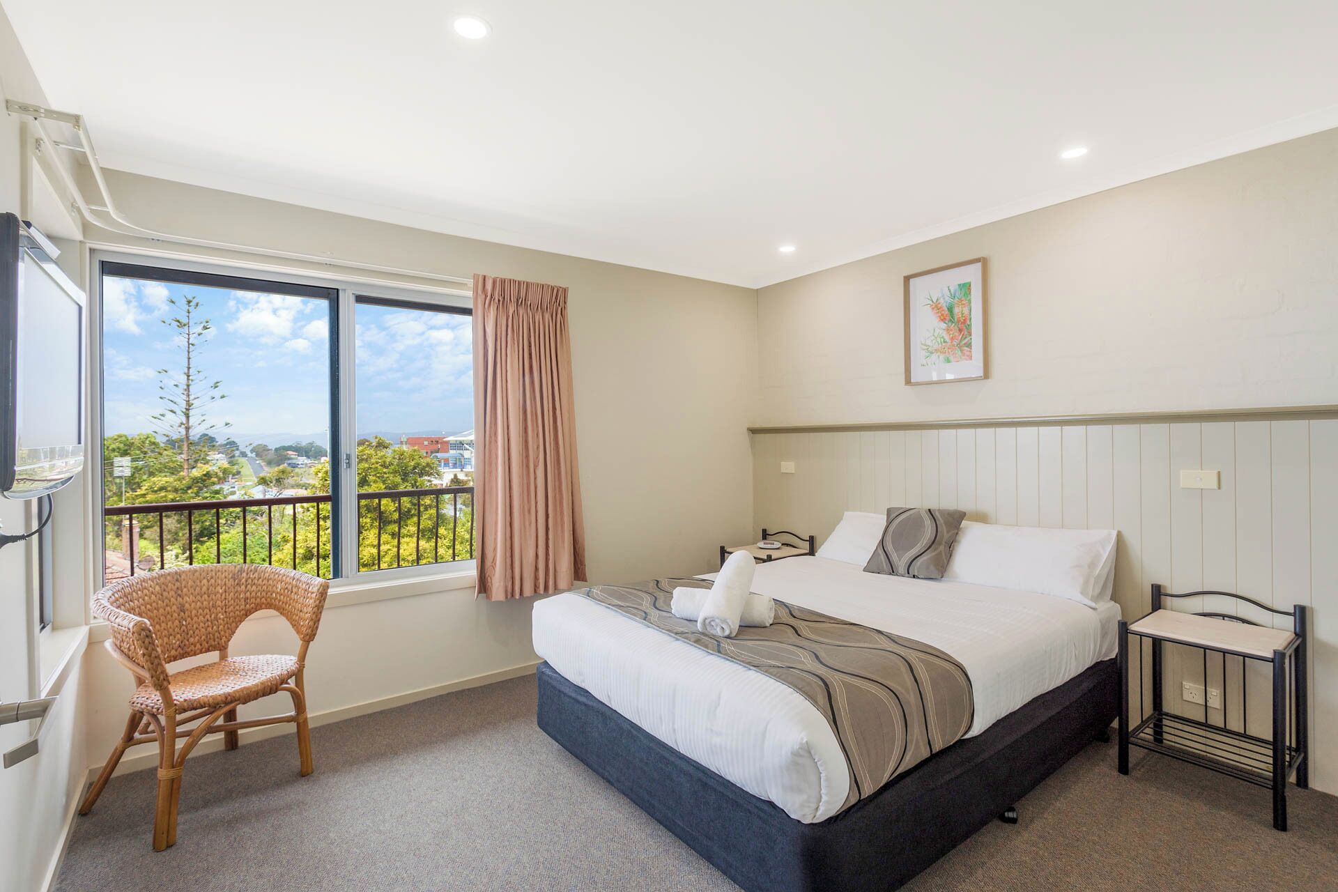 A bedroom with a bed, a chair, a bedside table, a window with a view, and a painting on the wall.