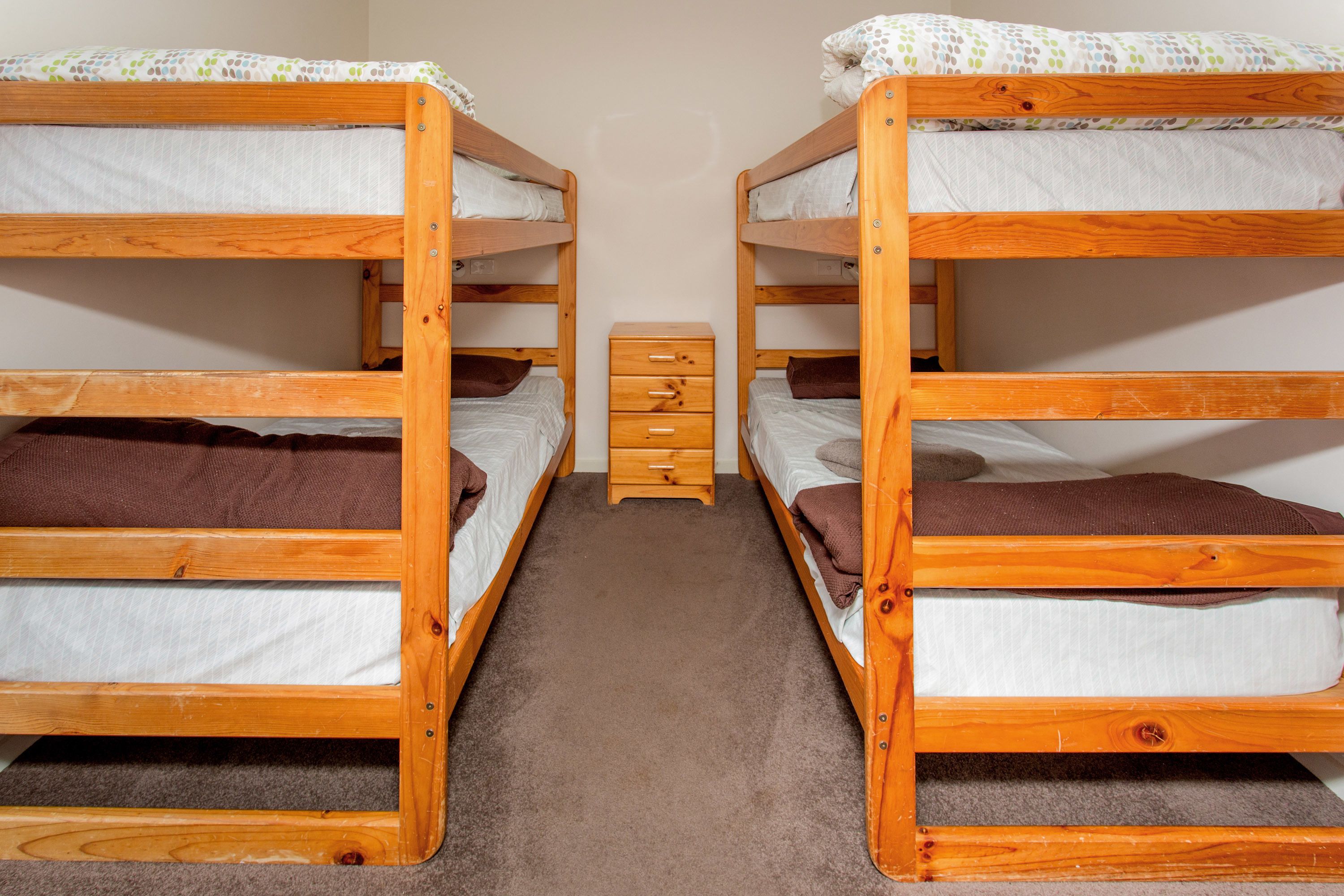 Een kamer met twee stapelbedden, elk voorzien van een kussen en dekbed, en een klein houten dressoir daartussen.