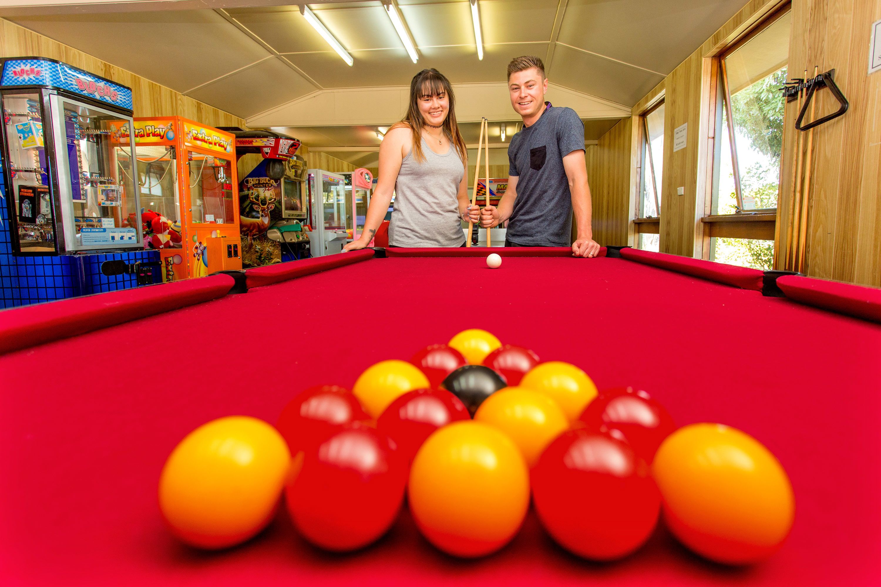 A pool table with balls, a man and a woman standing beside it, and arcade machines in the background.