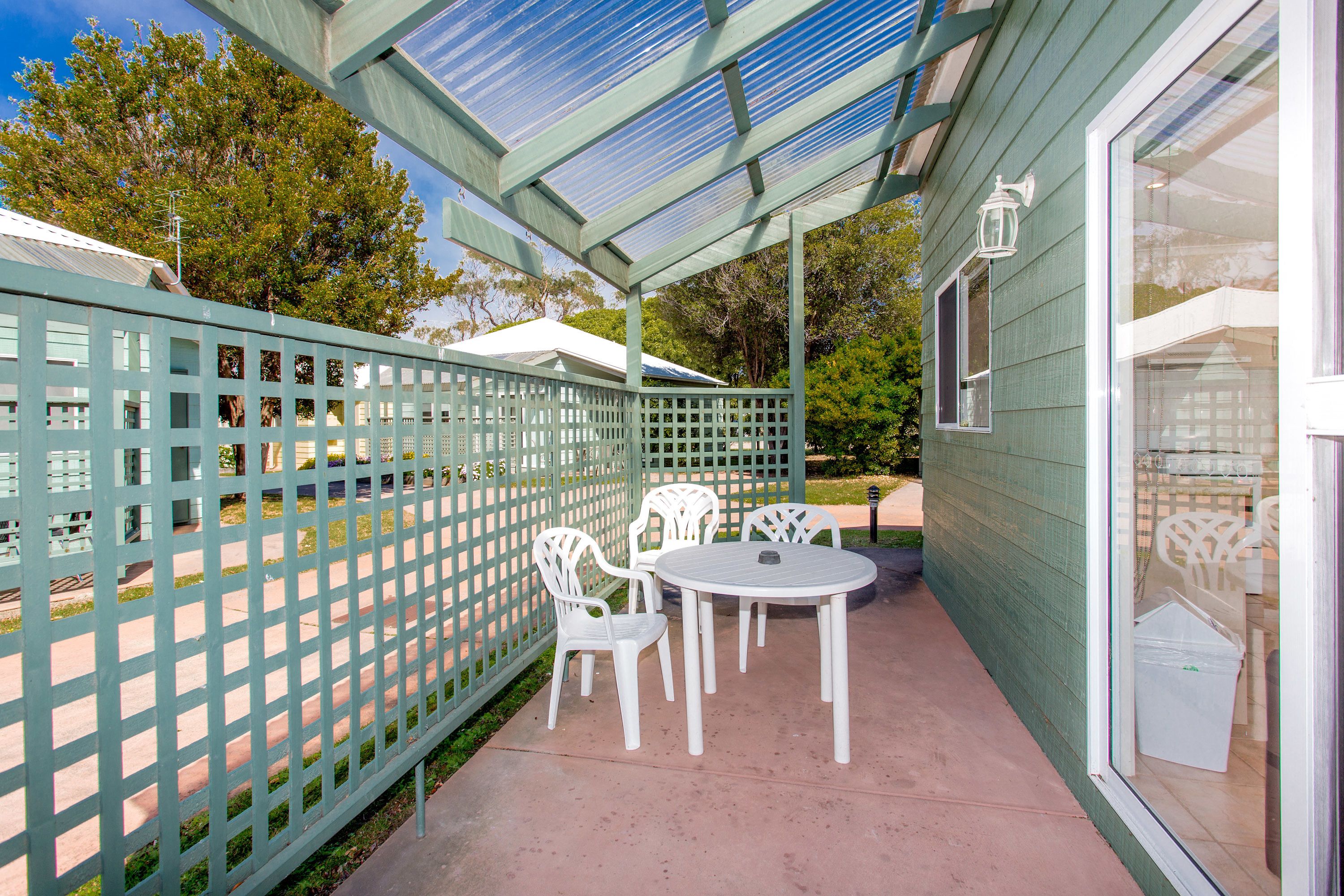 An outdoor patio with a table and chairs, a green fence, and a building with a window.