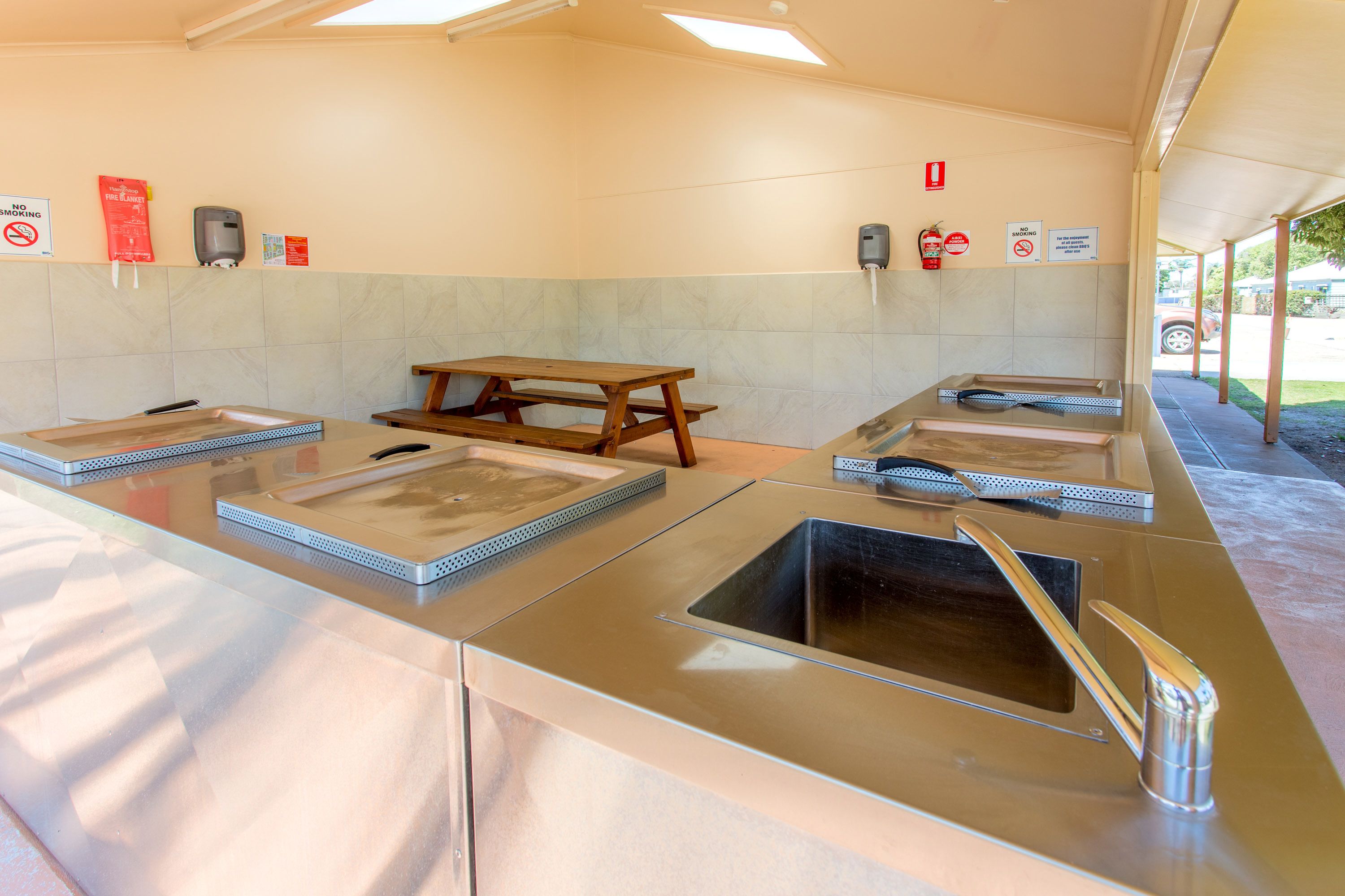 Un baño moderno y limpio con varios lavamanos, un banco de madera y carteles que indican prohibido fumar y consumir alimentos o bebidas.