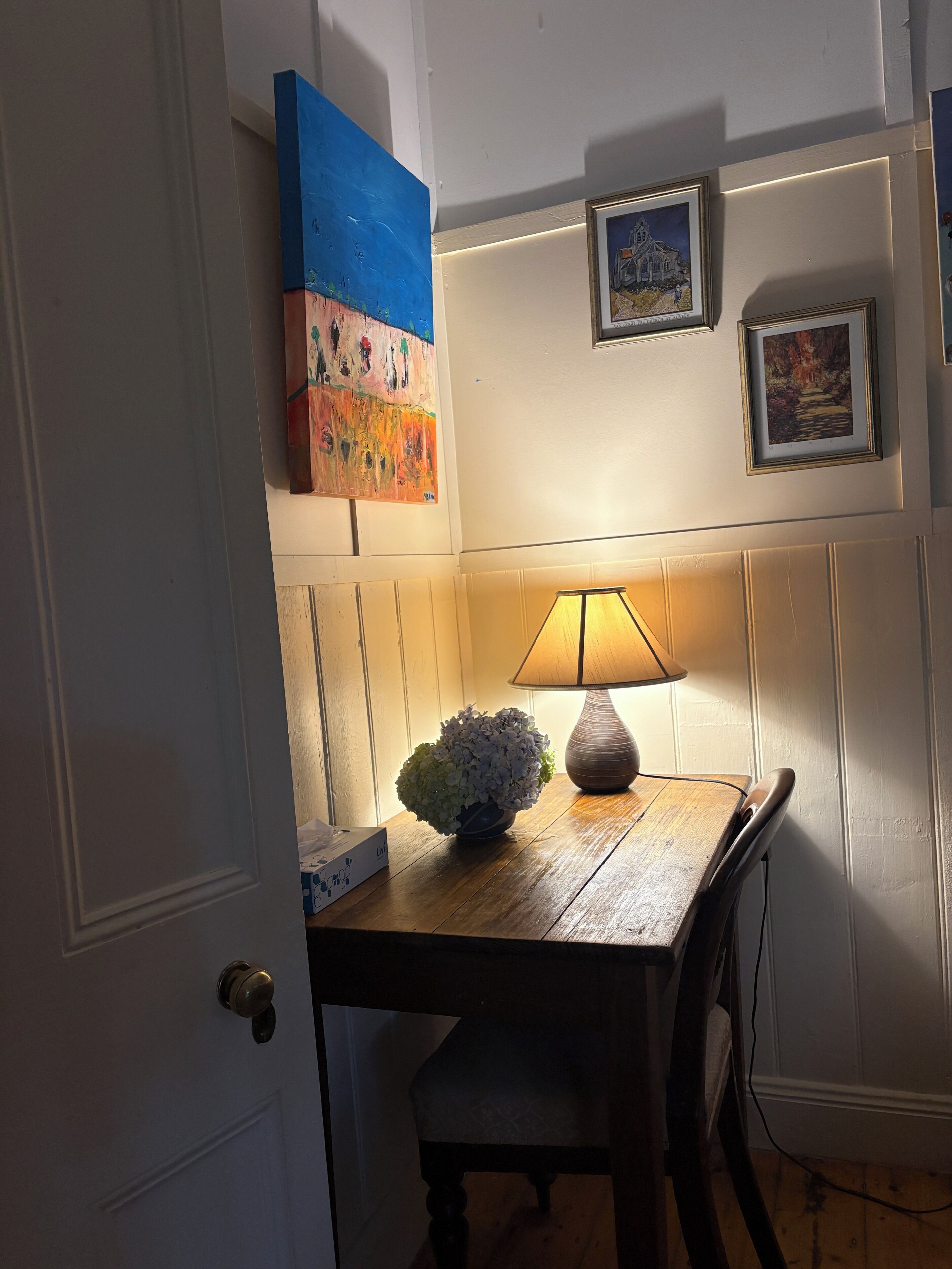 A room with a wooden table, a lamp, a chair, a door, and wall-mounted artwork.