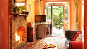 A cozy living room with a brick fireplace, a television, a sofa, and a dining area with a table set for two. - Como Cottages Accommodation (Olinda)