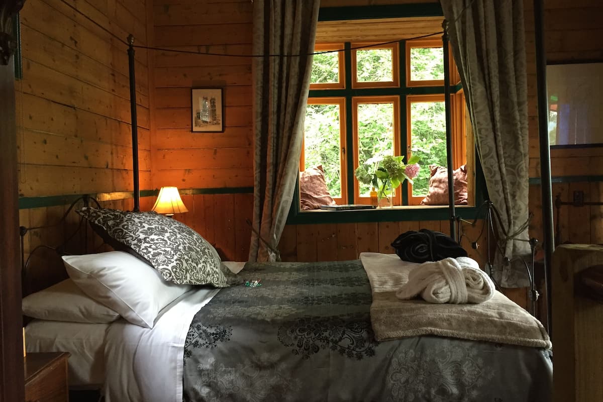 A wooden cabin bedroom with a bed, pillows, a bedside lamp, a window with curtains, and a view of greenery outside.
