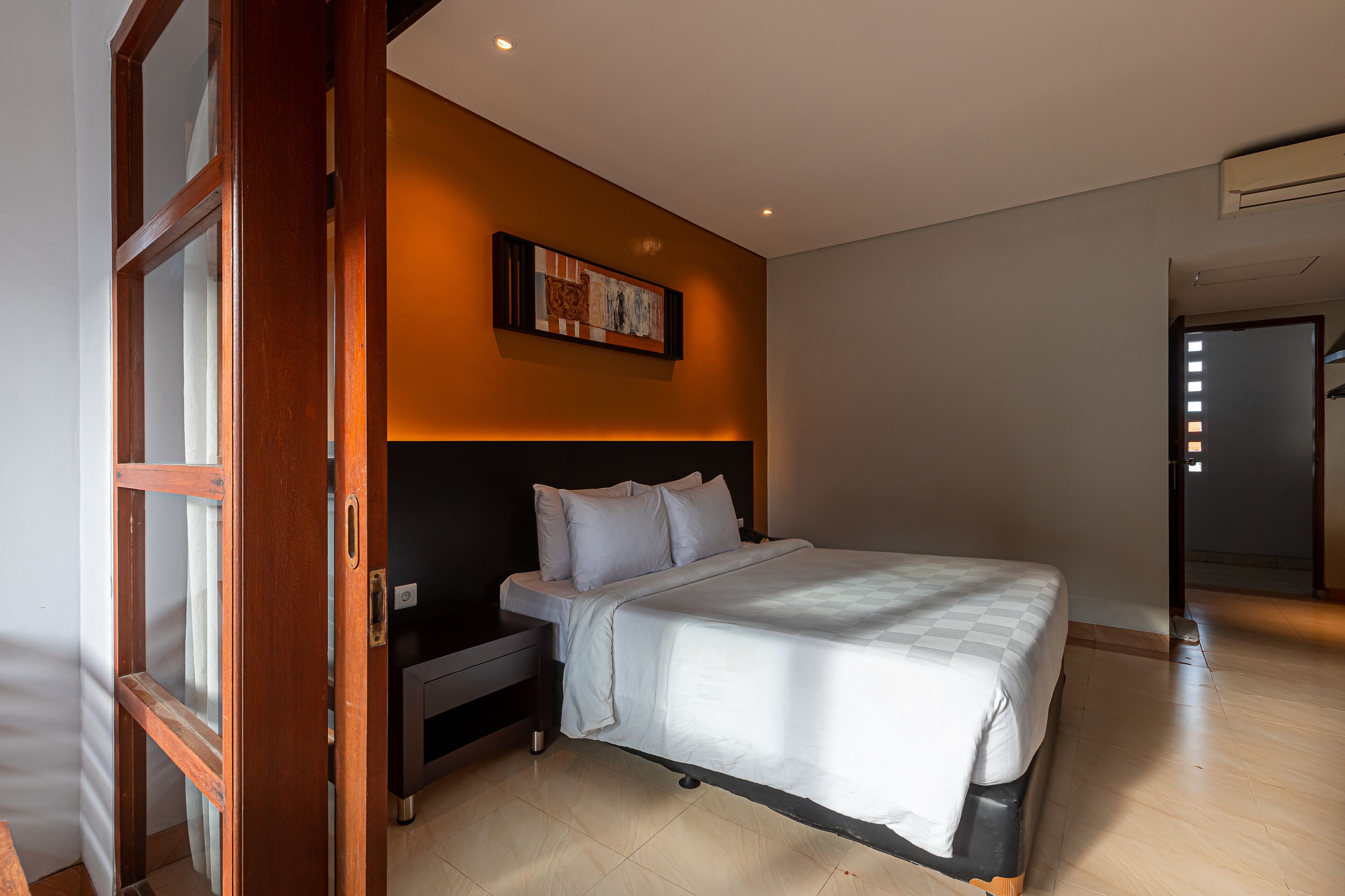 A bedroom with a bed, a nightstand, a wall-mounted artwork, and a door leading to another room.