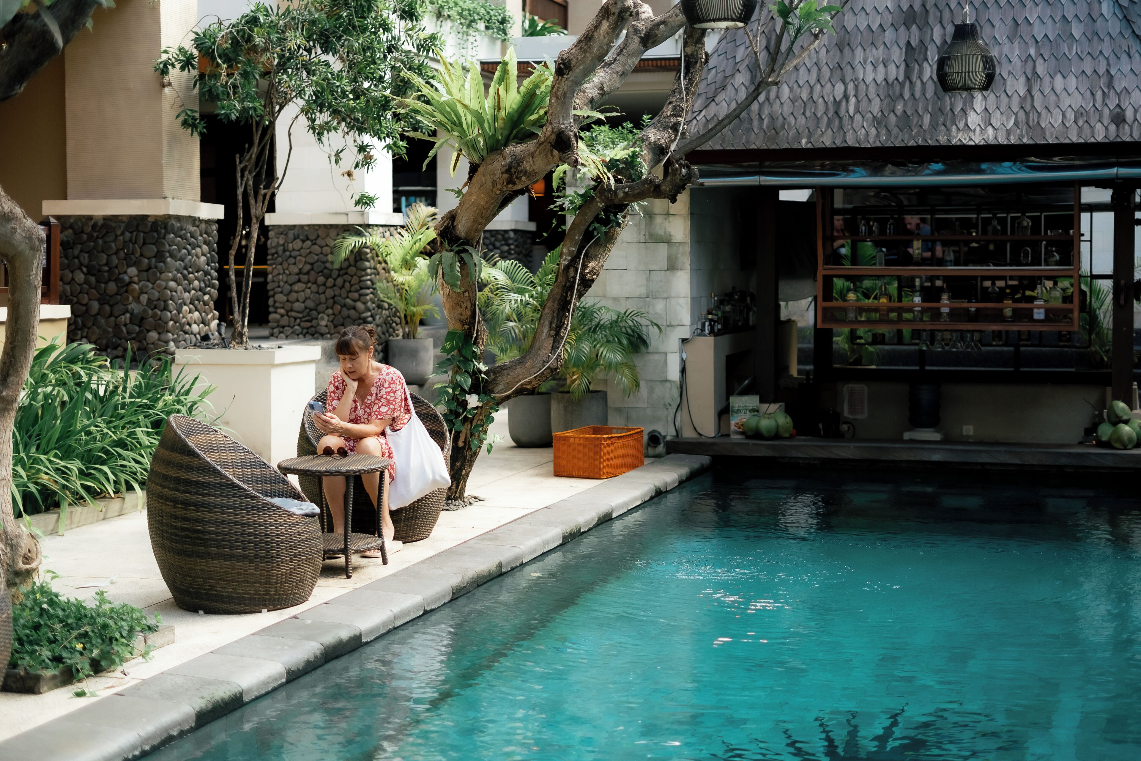 A poolside area with a woman sitting on a chair, surrounded by lush greenery and a stone wall.