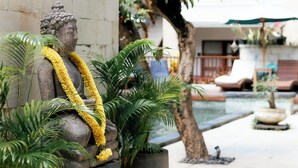 A statue of a seated figure with a yellow garland, surrounded by lush green plants and a stone wall. - Fourteen Roses Beach Hotel (Legian)