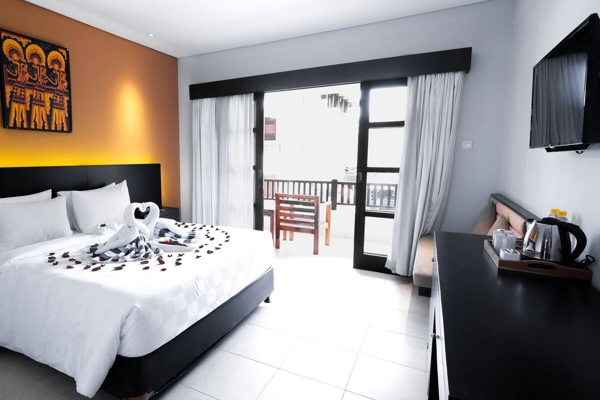 A hotel room with a large bed, a desk with a kettle, and a balcony with a chair.