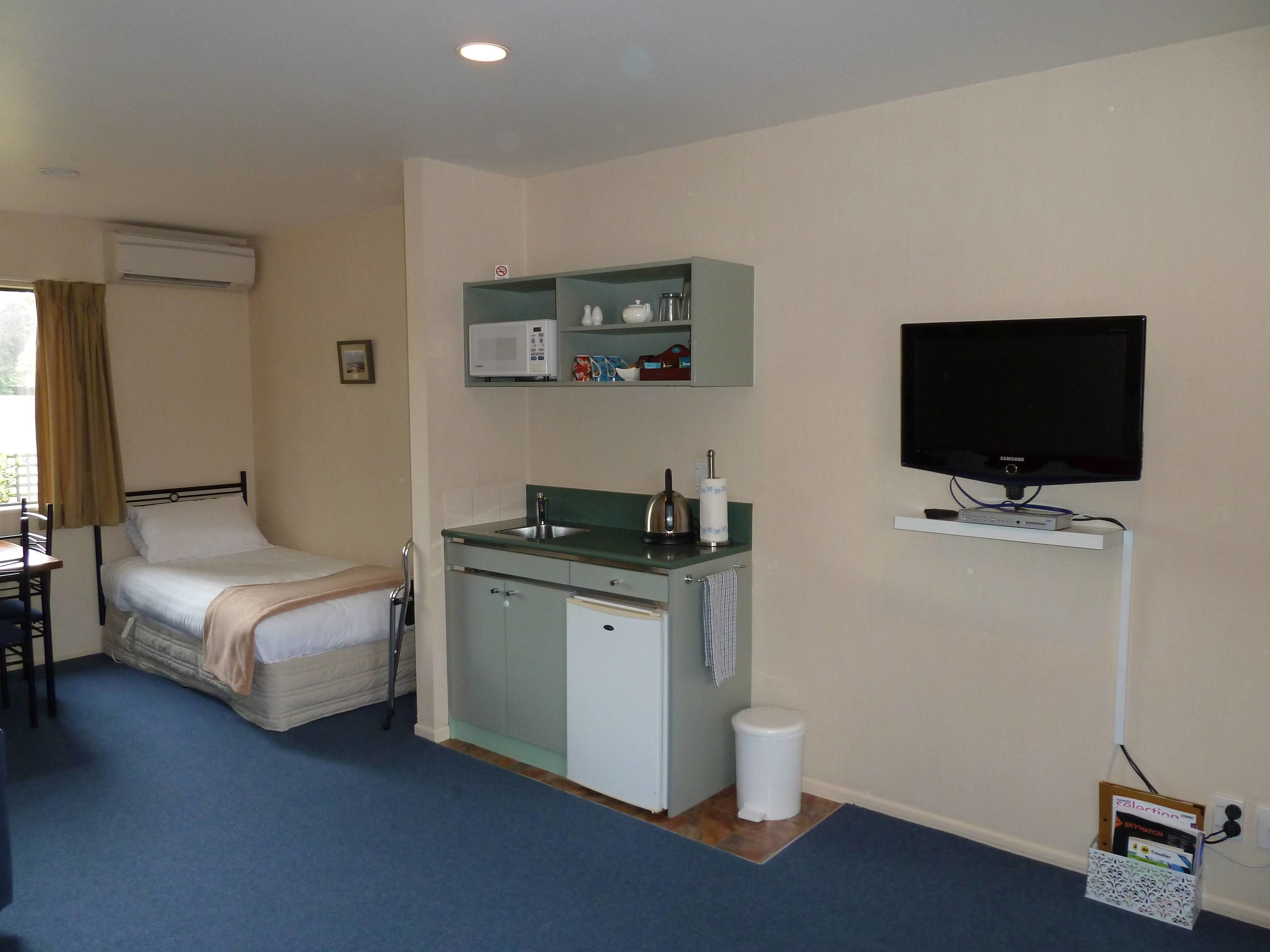 Habitación de hotel compacta con cama, kitchenette, televisor y un pequeño cubo de basura.