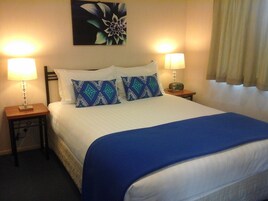 Quarto de hotel com uma cama, duas luminárias de cabeceira, um relógio e um quadro com flores na parede.