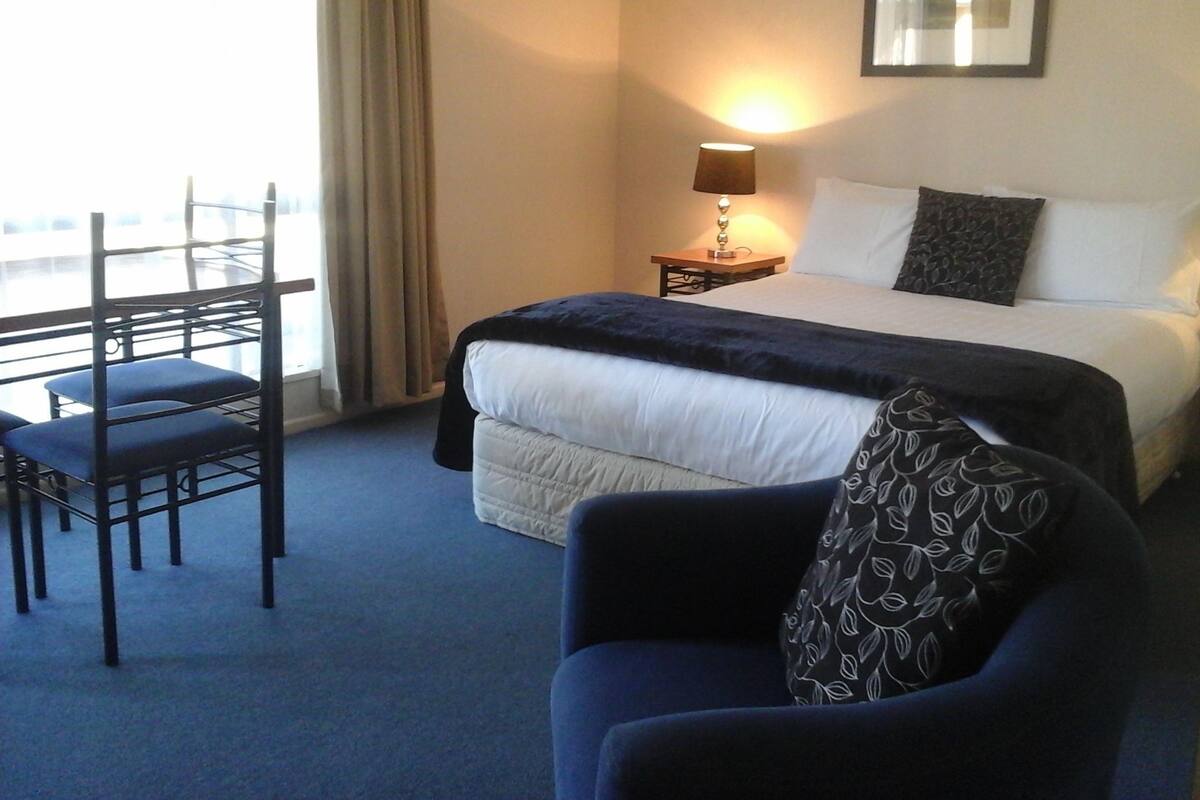 A hotel room with a bed, a chair, a nightstand with a lamp, and a window with curtains.