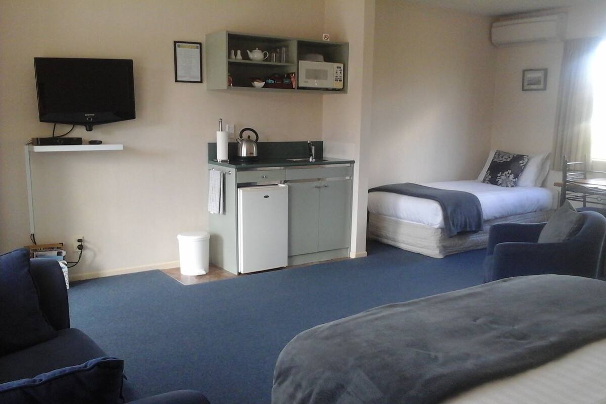 A compact hotel room with a bed, a small kitchenette, a television, and a desk area.