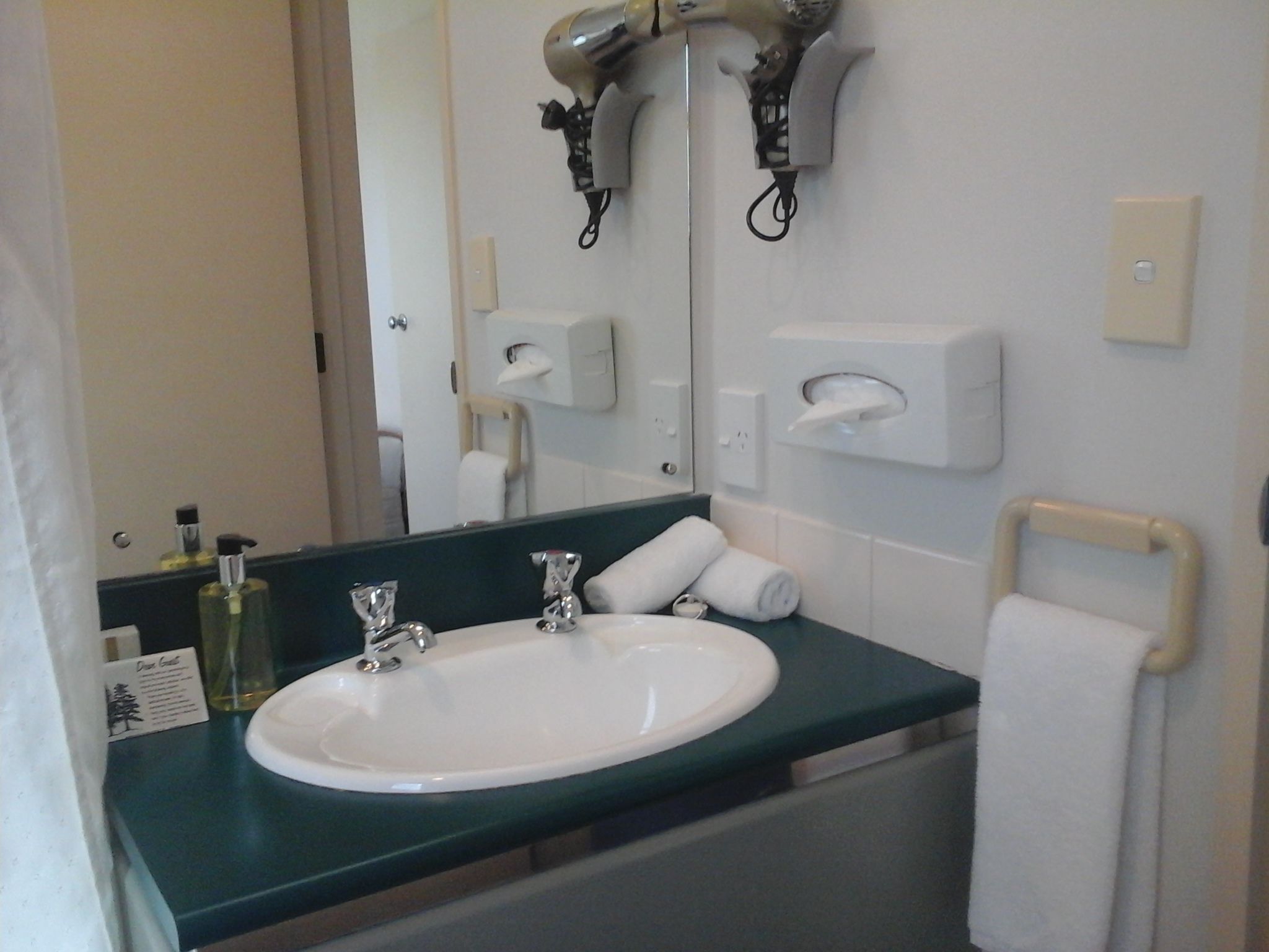 A bathroom with a white sink, two faucets, a mirror, a towel rack, and a hairdryer mounted on the wall.