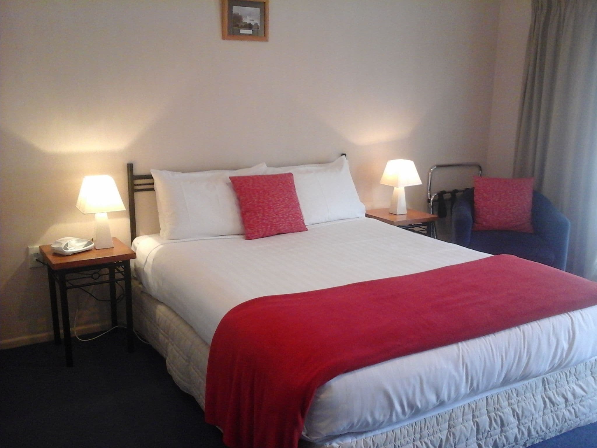 Una habitación de hotel con una cama grande, dos mesitas de noche, una silla y una mesita con una lámpara.